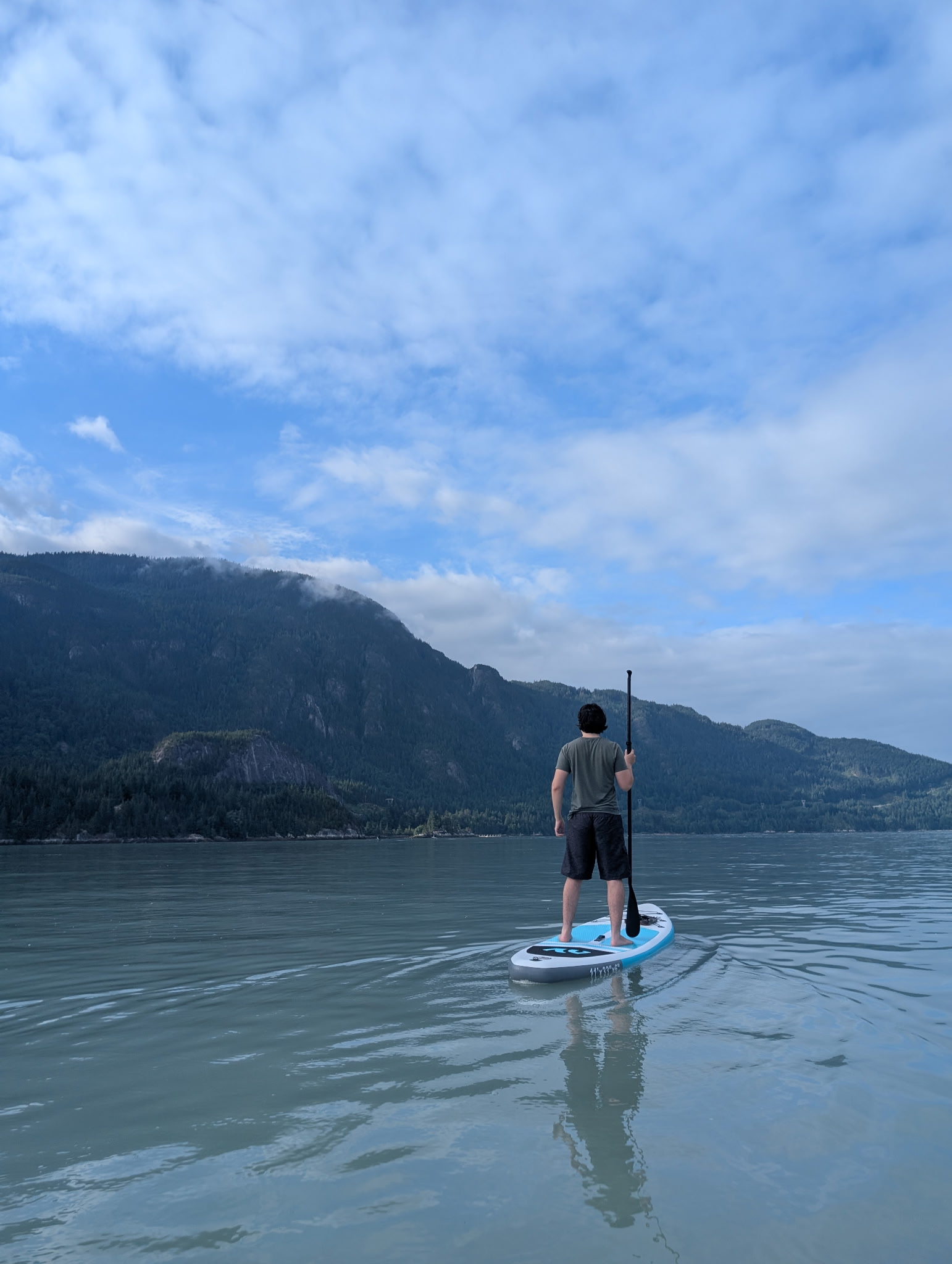 Paxton Cruz standing on a paddleboard on a still lake, facing forested mountains